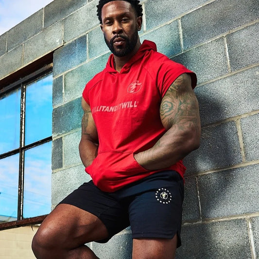 Man in red sleeveless hoodie and black shorts with logo leaning against brick wall.