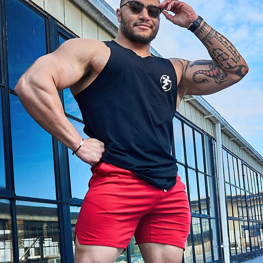 Man wearing black tank top and red athletic shorts. Tattooed arms, sunglasses.