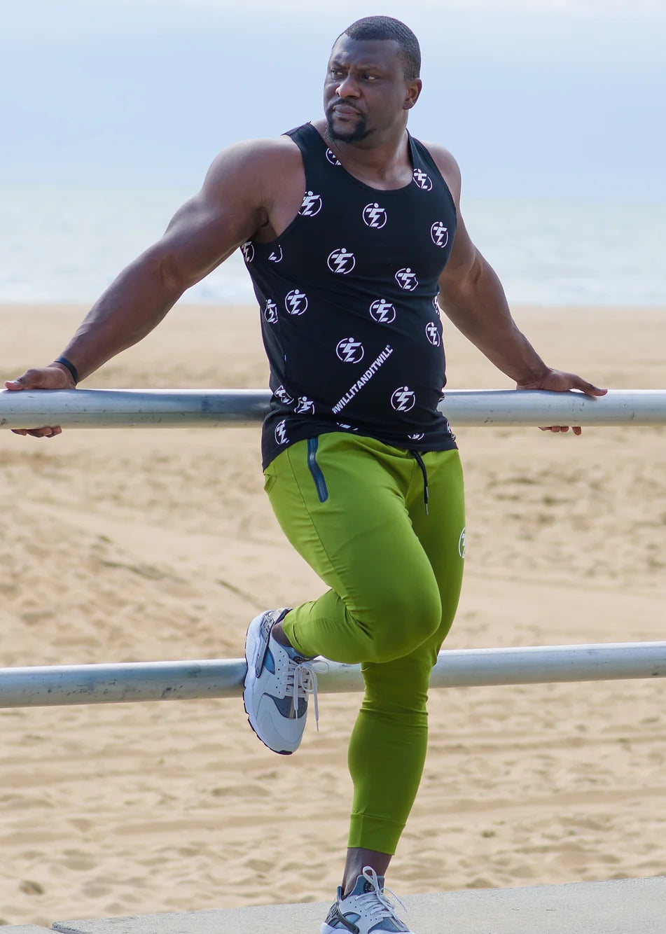 Man in black tank top and green joggers with white and grey sneakers on a beach background.
