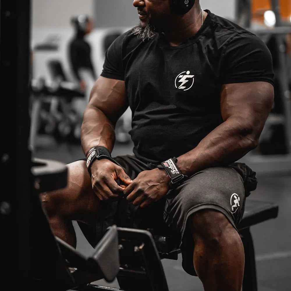 Man in black gym apparel (t-shirt and shorts) wearing wrist straps, seated at a gym.