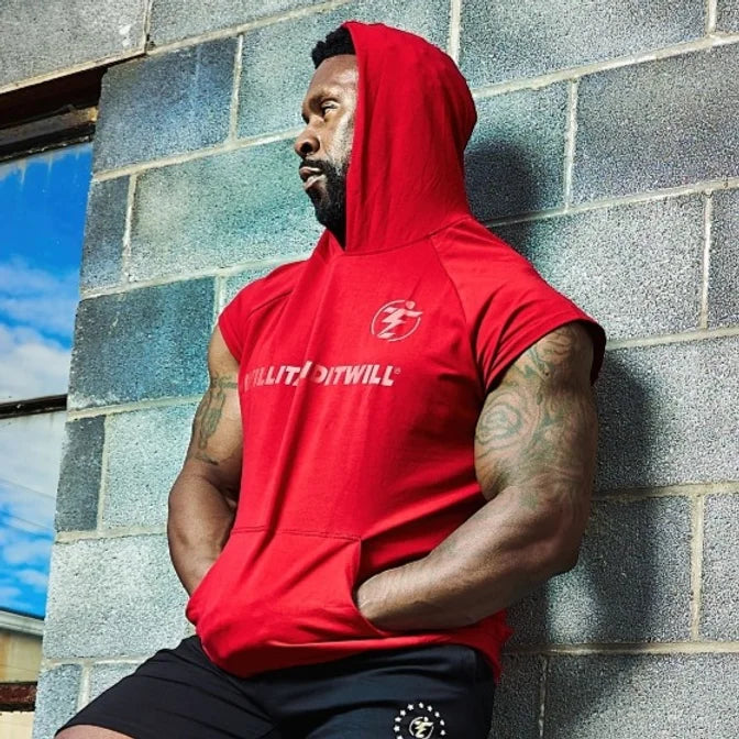 Man in red sleeveless hoodie and black shorts leaning against a brick wall.