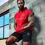 Man in red sleeveless hoodie and black shorts leaning against a brick wall.
