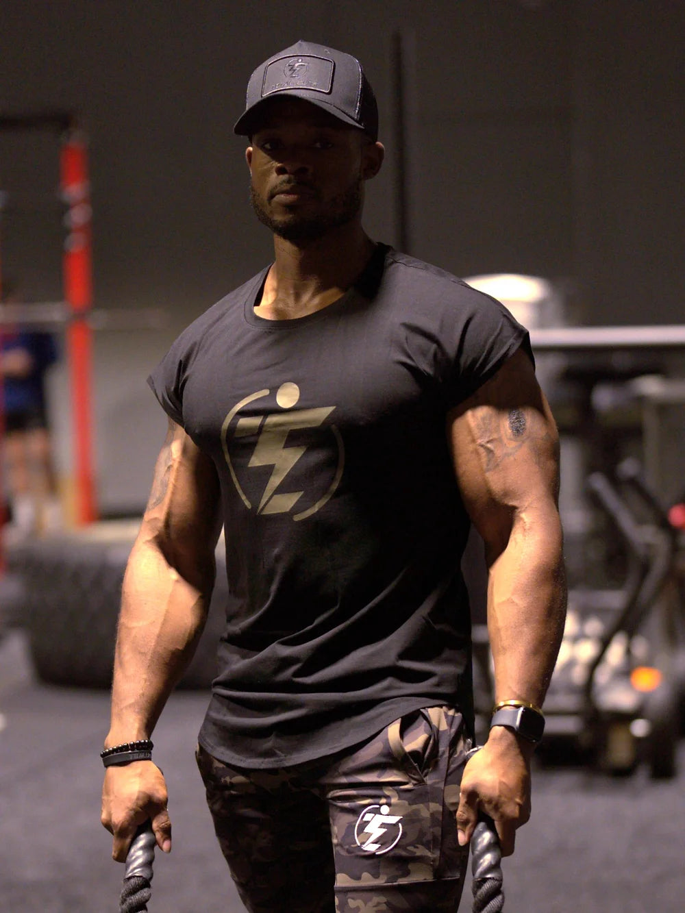 Man in black tee and camo shorts with logo holding battle ropes in a gym setting.