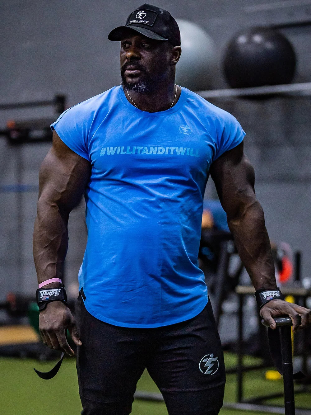 Man in blue #WILLITANDITWILL T-shirt and black athletic shorts with Total Elite logo at the gym.
