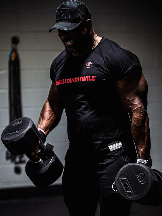 Man lifting dumbbells wearing a black WILLITANDITWILL t-shirt and hat in the gym.
