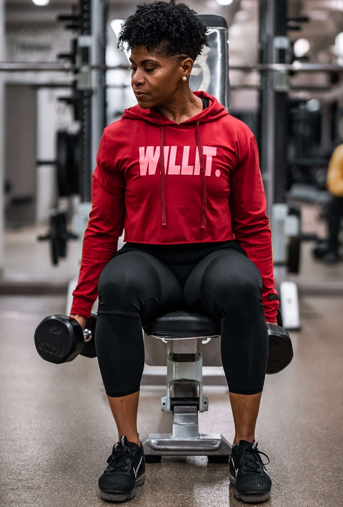 Woman lifting weights in a gym wearing a red WILLIT cropped hoodie and black leggings and shoes.