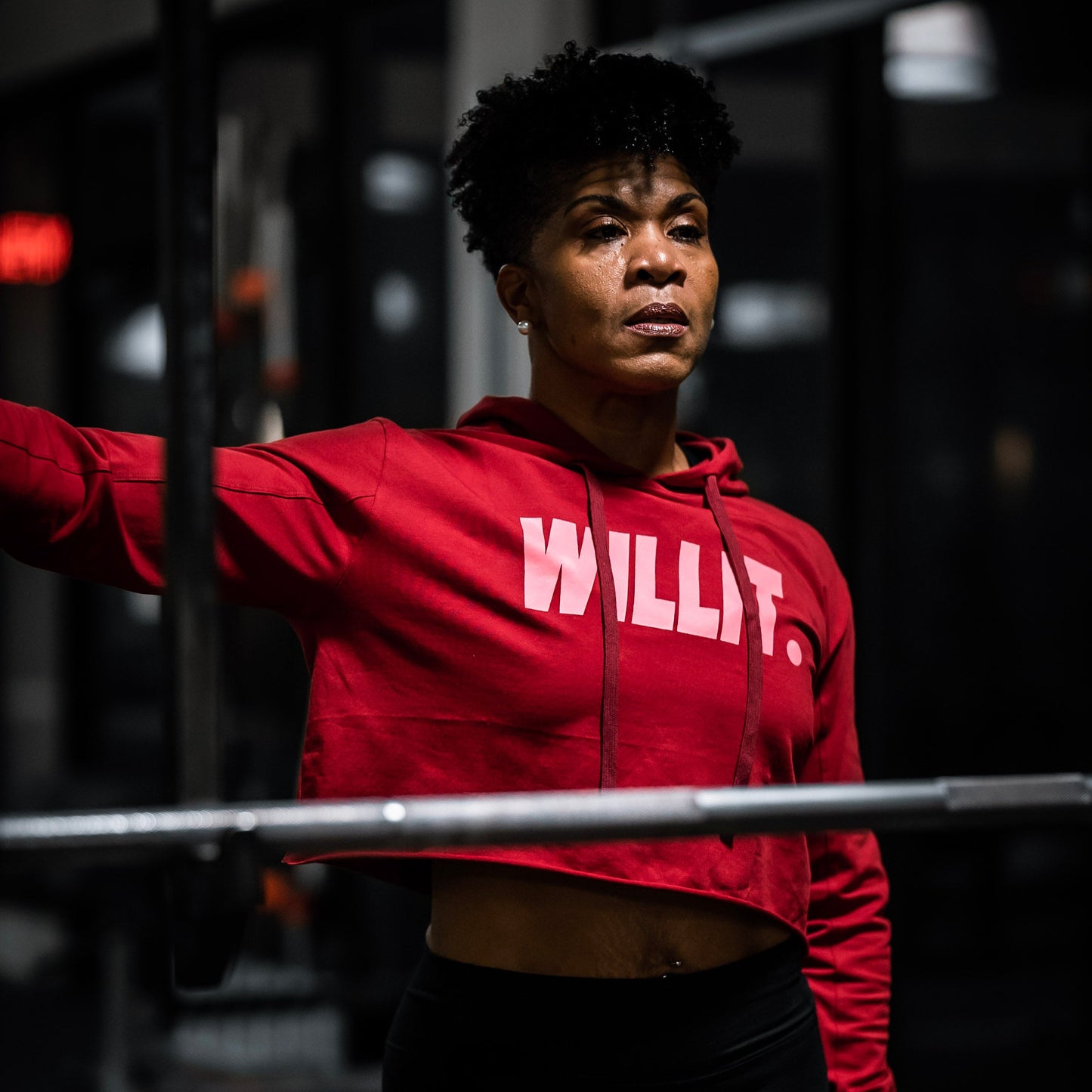 Woman in red 'WILLIT.' cropped hoodie at the gym with a barbell. Activewear fashion.