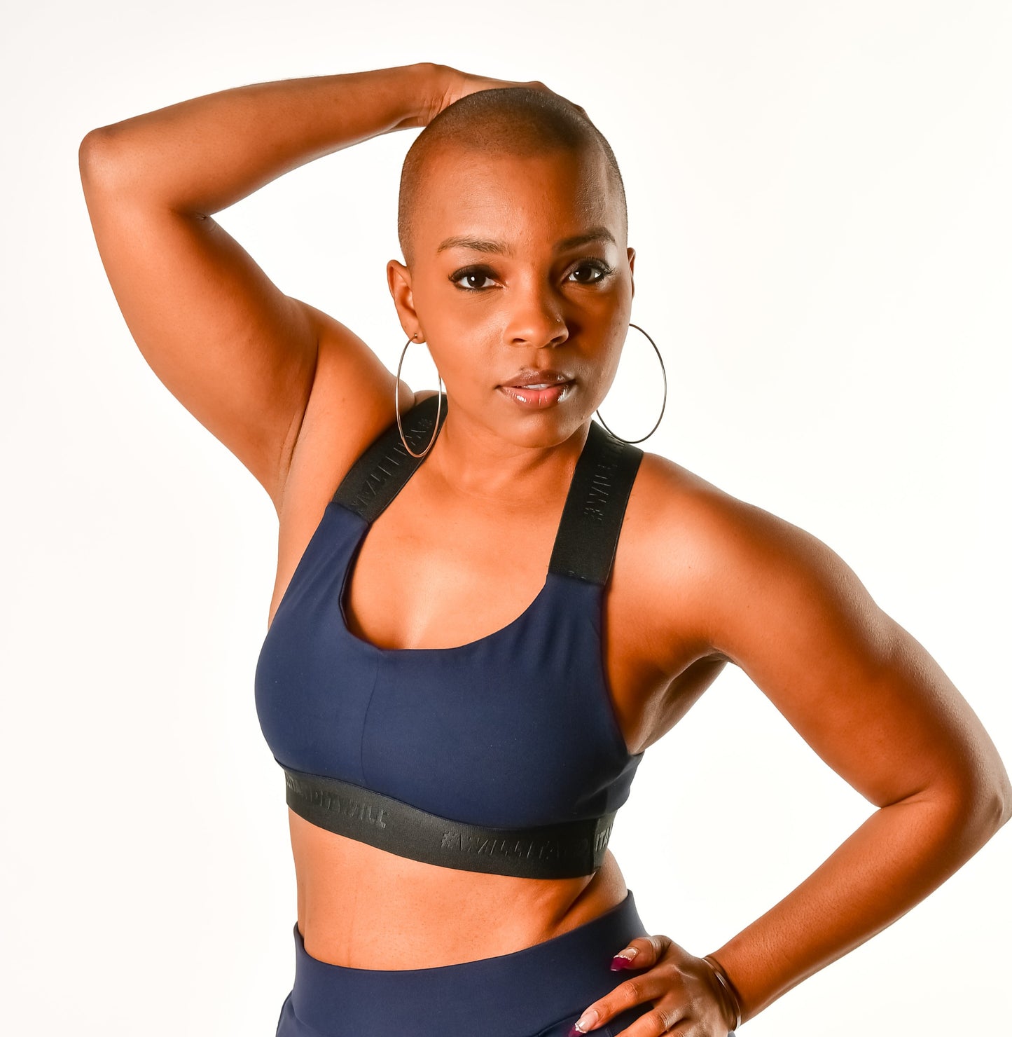 Woman in navy blue sports bra and leggings, posing with hand on head. Hoop earrings visible.