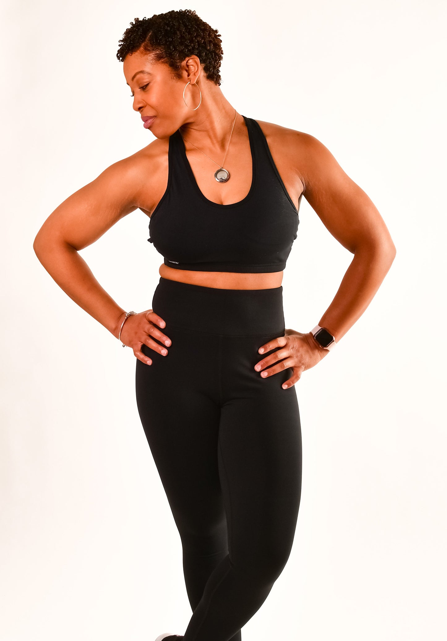 Woman in black sports bra and leggings with watch and necklace posing on white background.