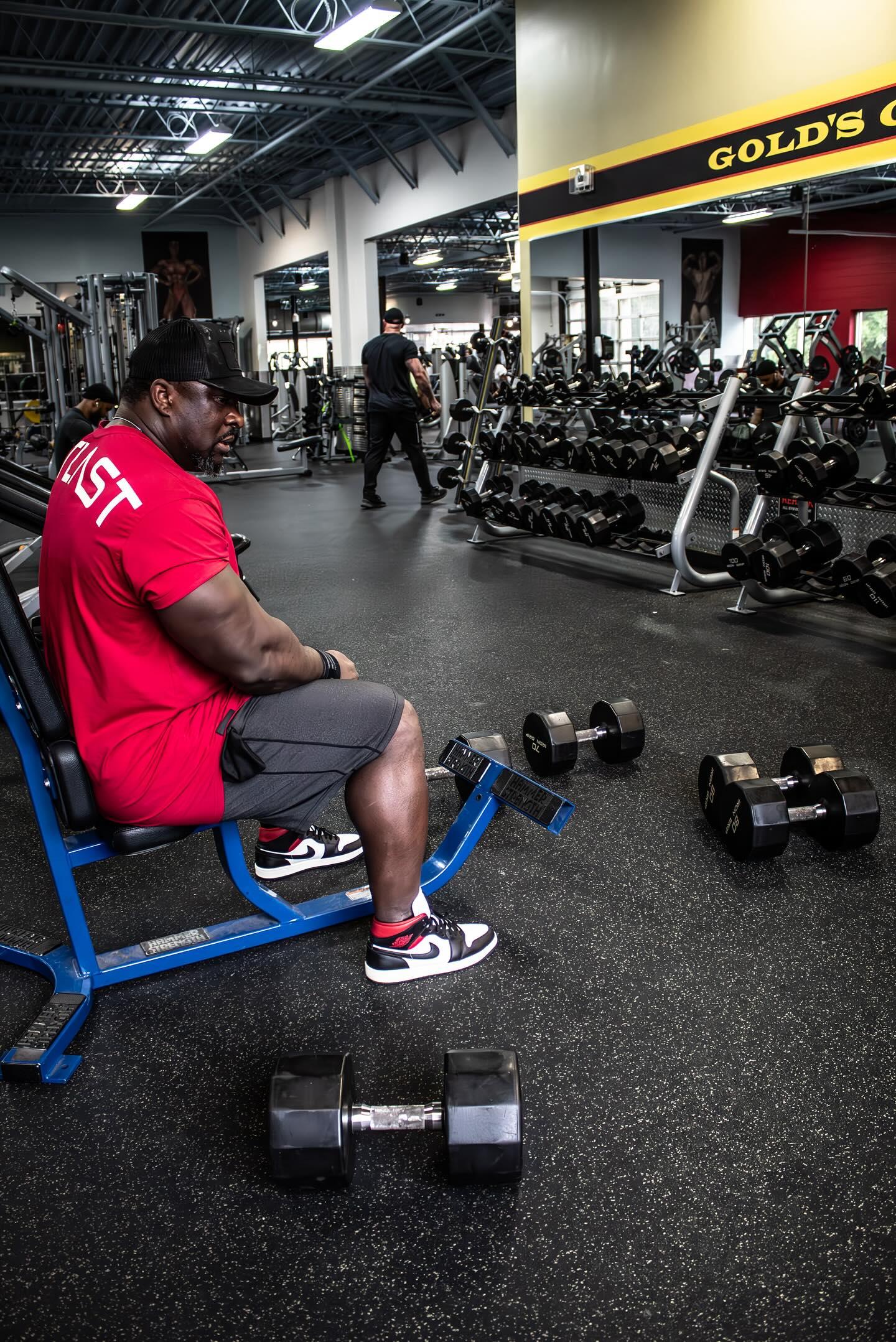 Man resting between sets with dumbbells at Gold's Gym, wearing red shirt, gray shorts and Air Jordans.