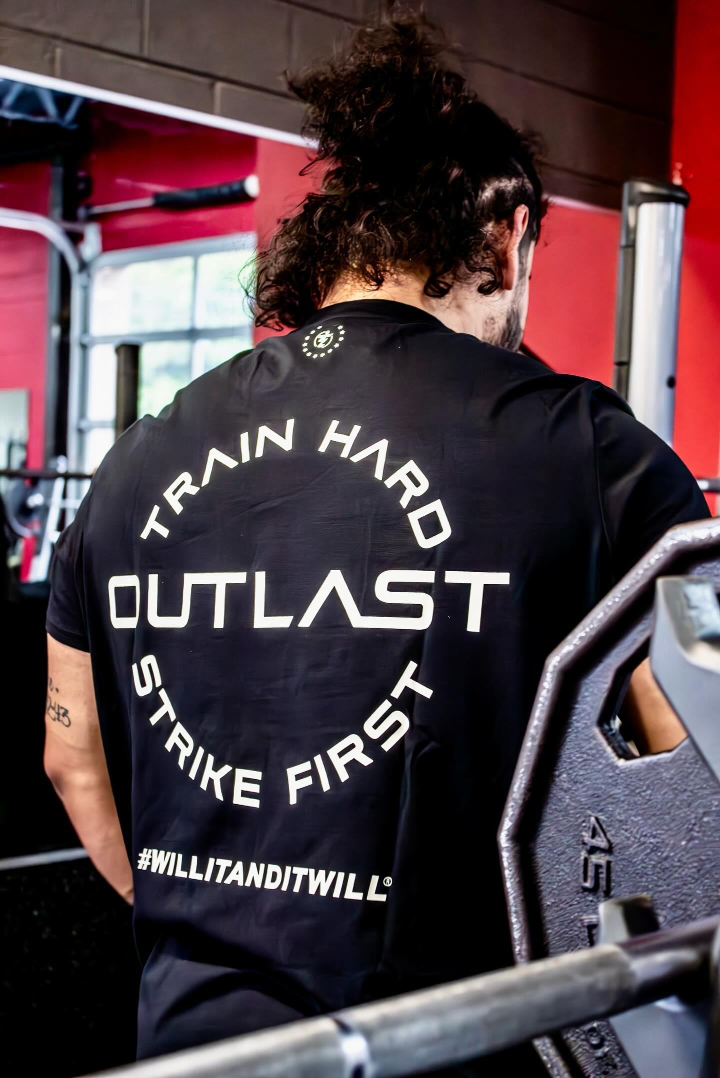 Black workout shirt with 'Train Hard, Outlast, Strike First' text and #WILLITANDITWILL slogan in gym.