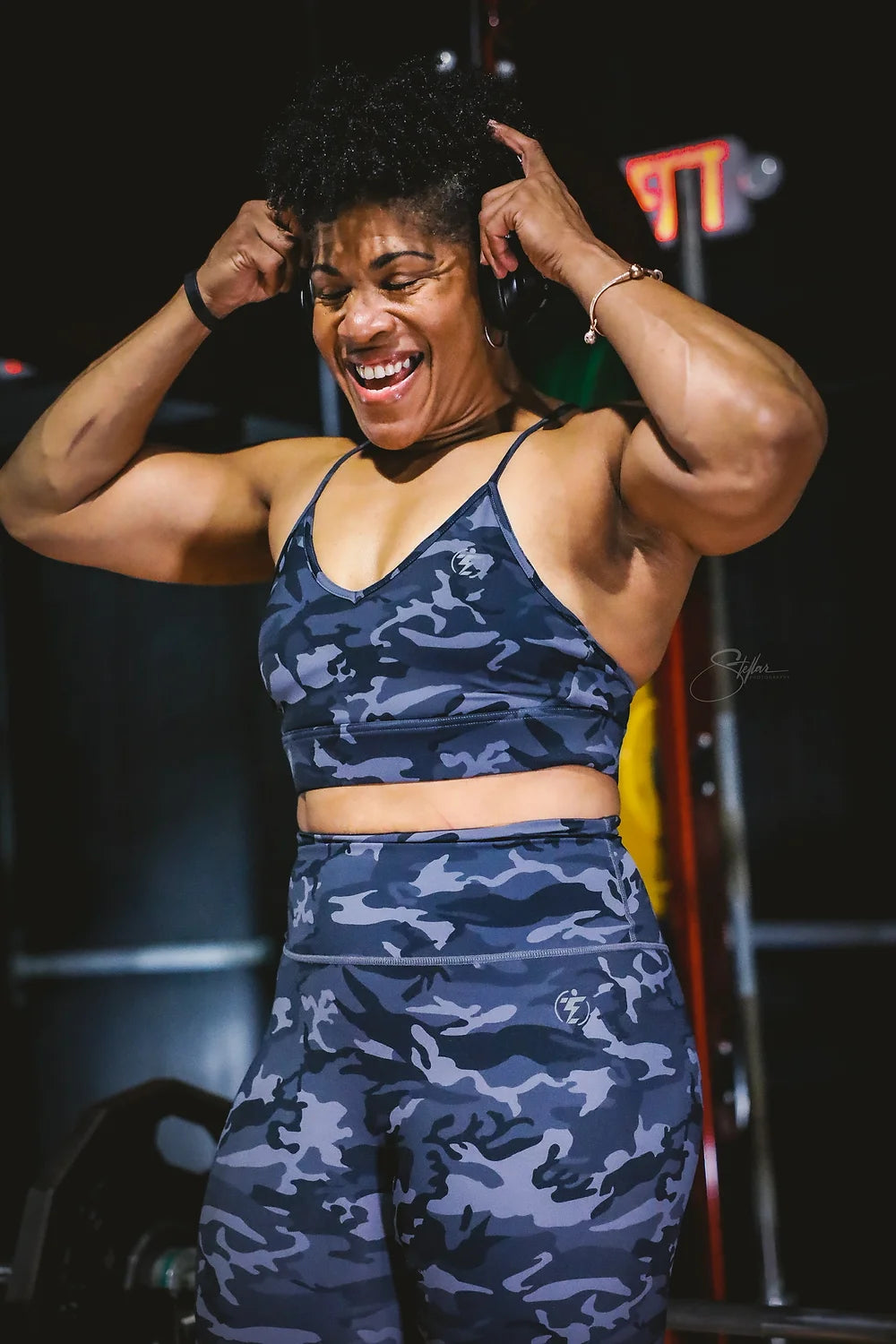 Woman in camo print workout set (sports bra and leggings) at the gym, smiling with headphones on.