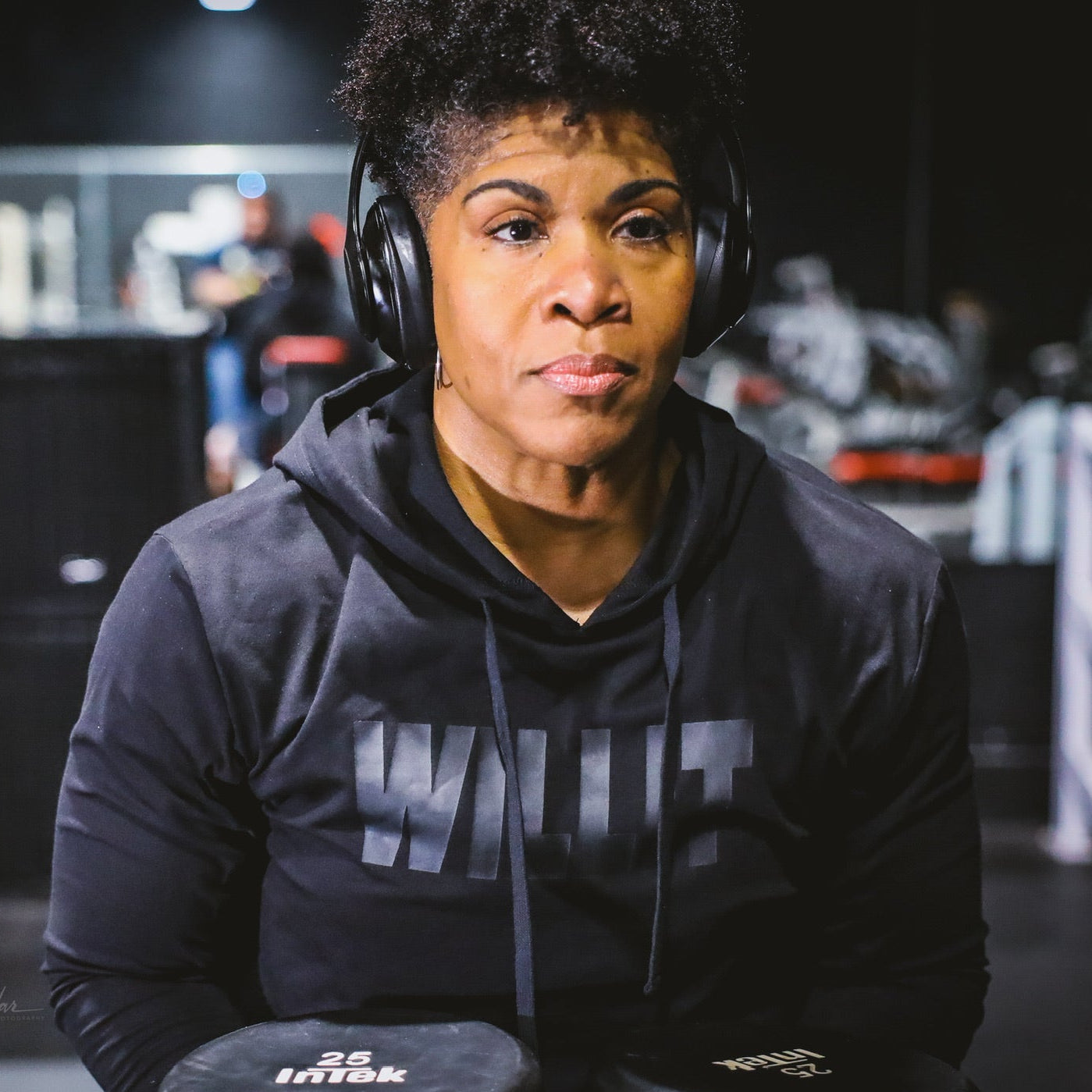 Woman with headphones wearing a Wulfit hoodie holding 25lb Intek dumbbells at the gym.