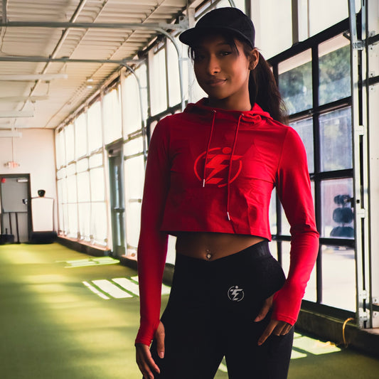 Woman wearing red cropped hoodie and black leggings with logo in gym setting.