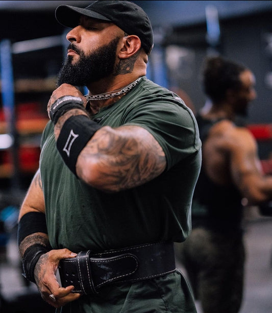 Muscular man with tattoos, weightlifting belt, wrist wraps, and a chain necklace in a gym.