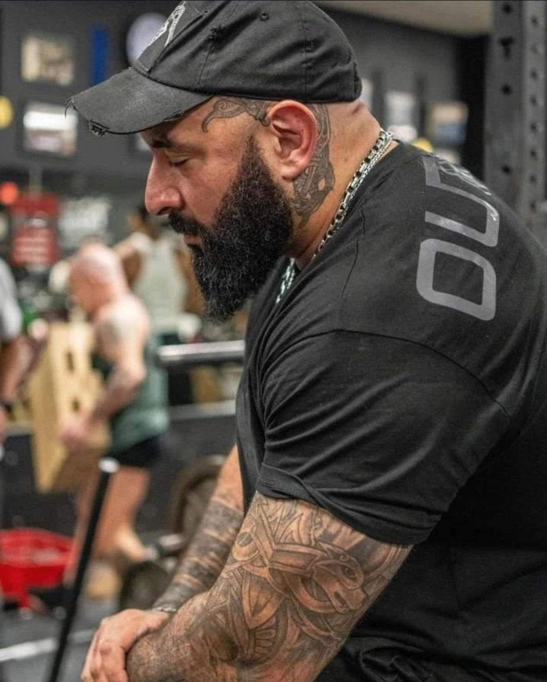 Man with tattoos wearing a black t-shirt and cap in a gym.