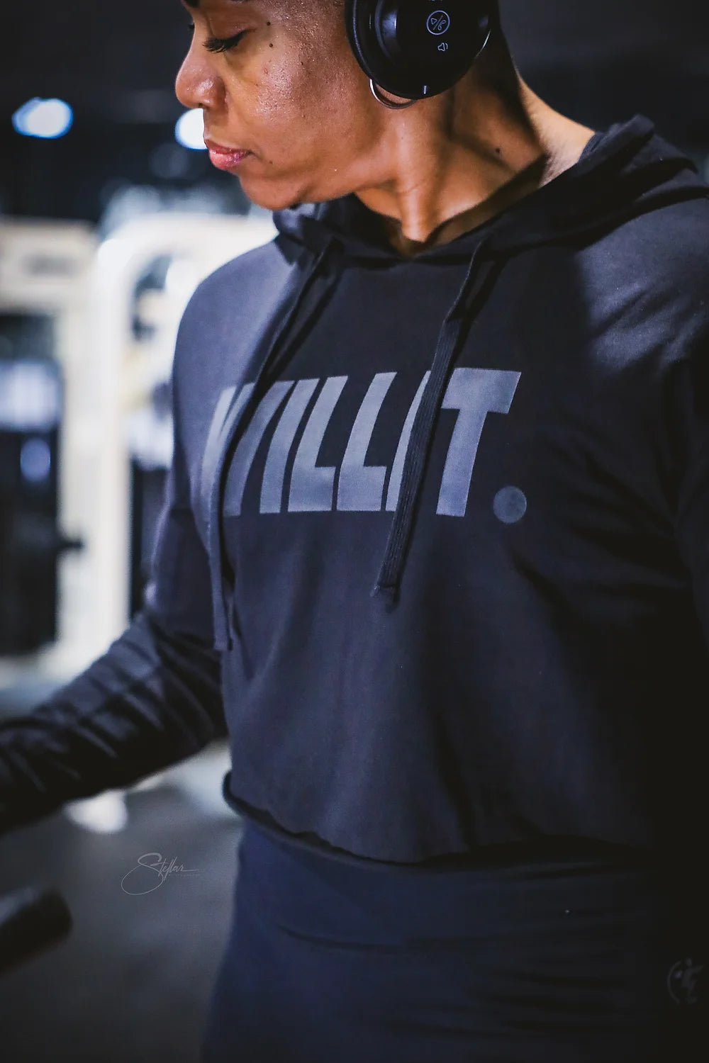 Woman in a dark KILLIT cropped hoodie and matching leggings at the gym wearing black wireless headphones.