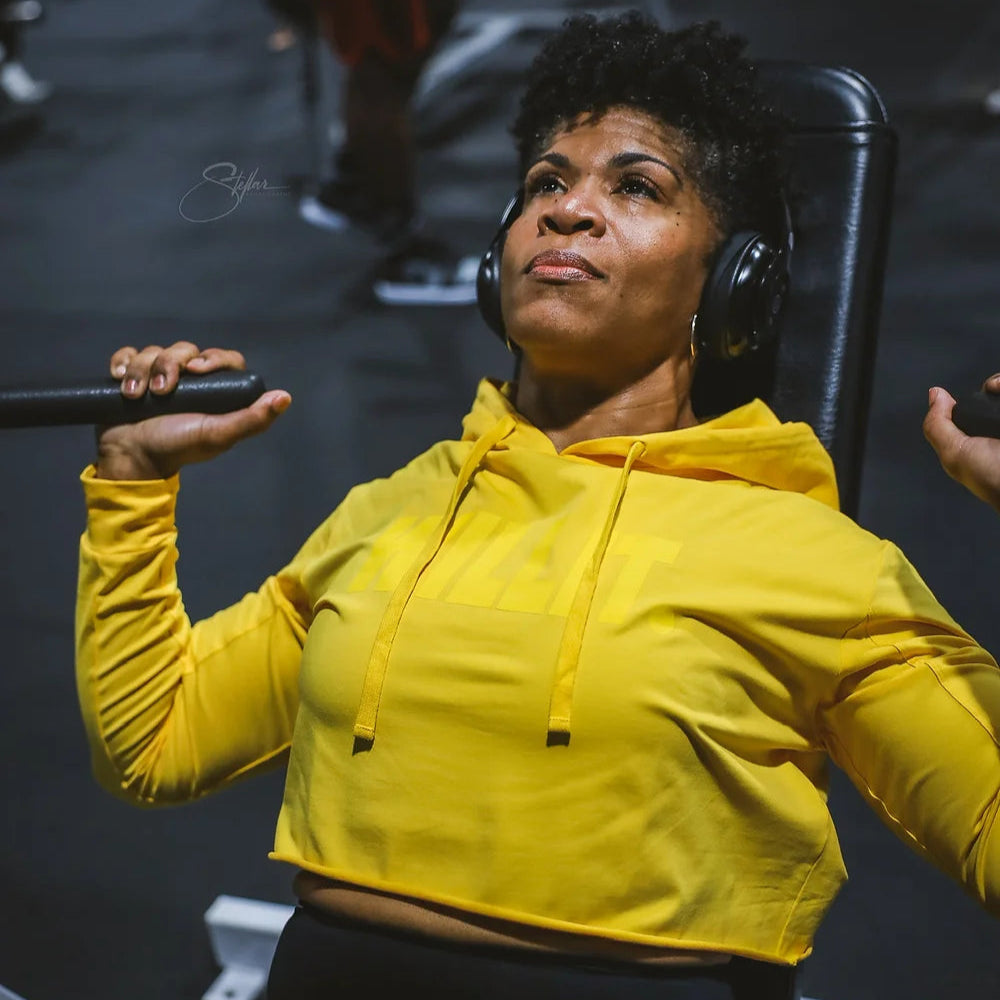 Woman using gym equipment wearing a yellow cropped hoodie and headphones.