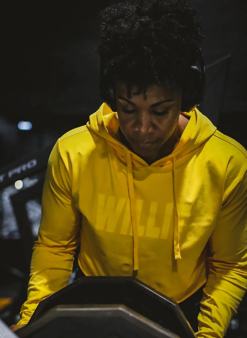 Woman in yellow hoodie with headphones using treadmill for indoor workout. Hoodie has "WALLIE" text.