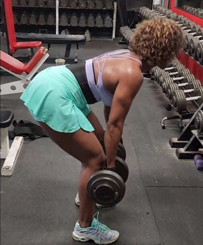 Woman lifting dumbbells in gym wearing waist trainer, sports bra and tennis skirt.